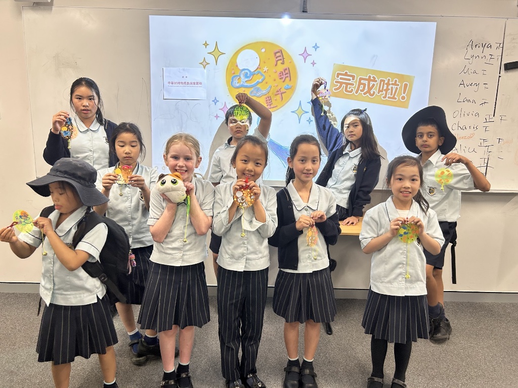 學生於「月明千里」多媒體螢幕前高舉手作掛飾，展現結合漢字成語與手工編織的學習成果。